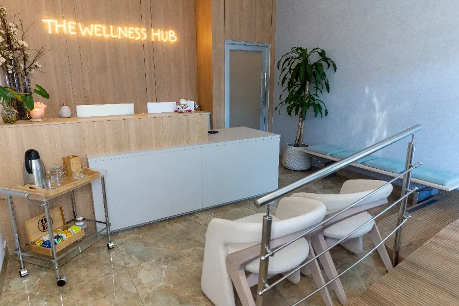 Una moderna área de recepción o sala de espera con un mostrador de madera clara y un letrero de neón que dice "THE WELLNESS HUB". Hay una estación de té y café en un carrito, una planta grande, un banco de madera con cojines azules y sillas tapizadas de color crema, con un pasamanos metálico en primer plano sobre un suelo de baldosas de mármol.